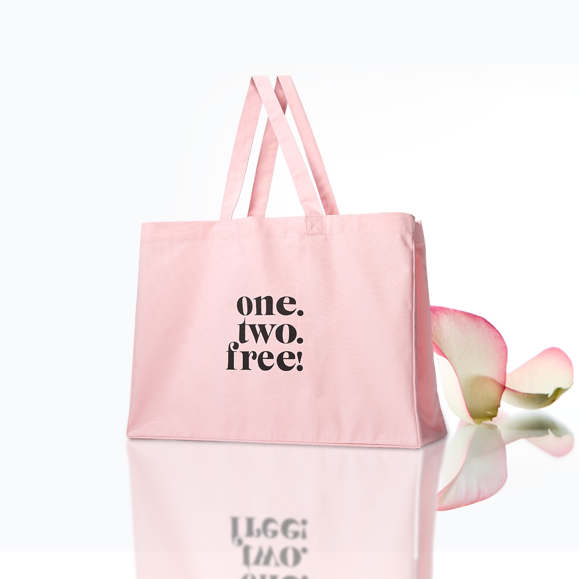 Eine hellrosa Stofftasche mit der Aufschrift „one. two. free!“ in schwarzer Schrift, daneben liegt ein rosafarbenes Blütenblatt.