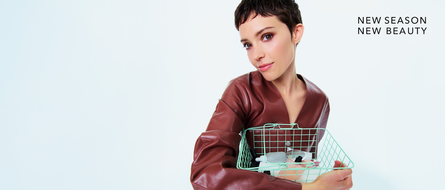 Eine Frau mit kurzen braunen Haaren und einem braunen Oberteil hält einen mintgrünen Korb mit verschiedenen Beauty-Produkten in der Hand, daneben steht der Text "NEW SEASON NEW BEAUTY".