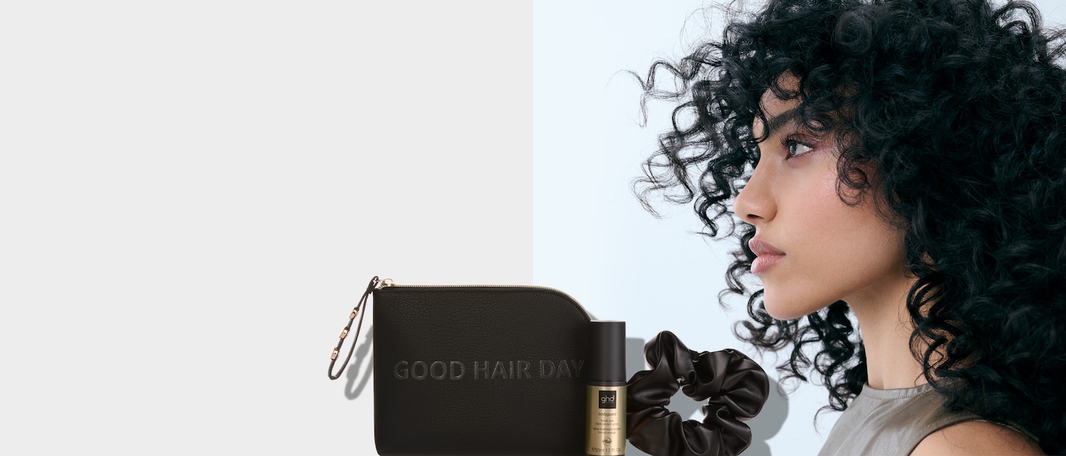Das Bild zeigt eine Frau mit lockigem Haar, die eine schwarze Tasche mit der Aufschrift „GOOD HAIR DAY“, ein ghd Hitzeschutzspray und ein schwarzes Haargummi in Satinoptik präsentiert.
