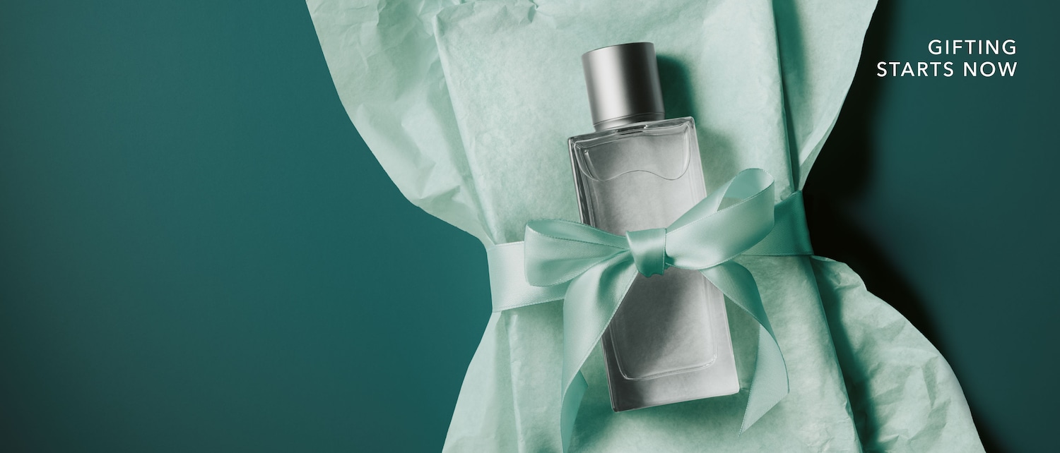 Ein eleganter Parfumflakon mit silberner Kappe liegt auf mintgrünem Seidenpapier, verziert mit einer passenden Schleife, während der Schriftzug Gifting Starts Now zum Schenken einlädt.