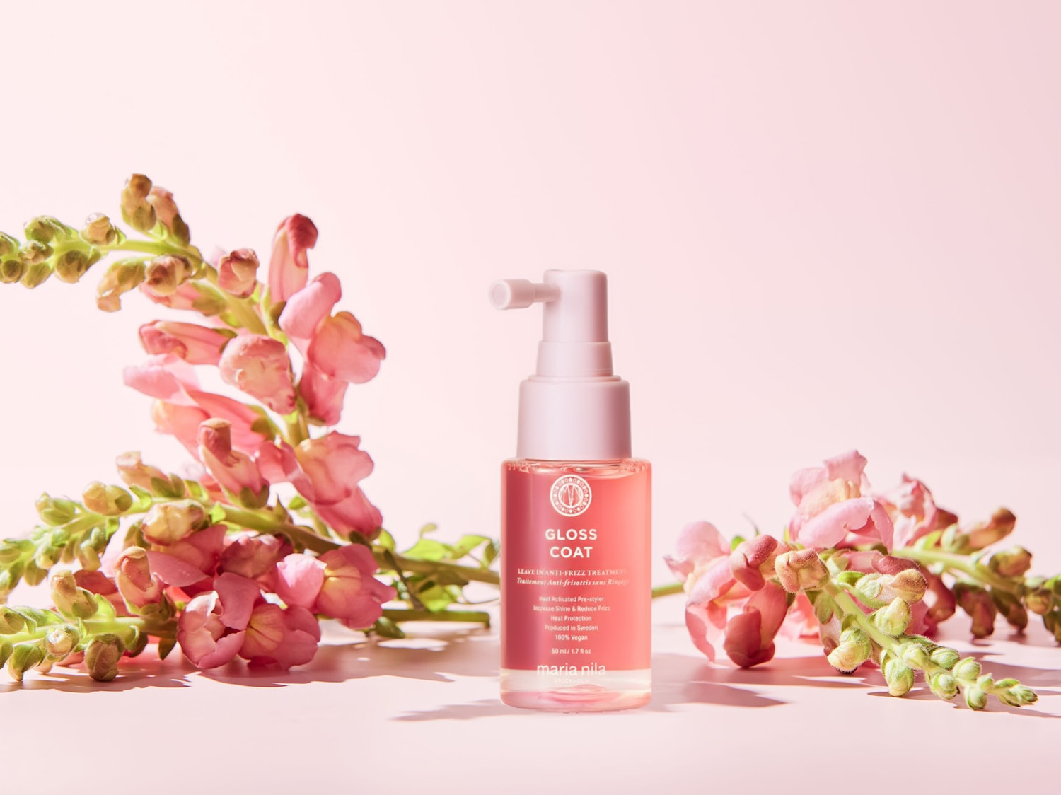 Das Maria Nila Gloss Coat Leave-In Anti-Frizz Treatment in einer 50-ml-Flasche mit rosa Flüssigkeit und weißem Pumpspender, umgeben von rosa Blüten, verspricht Glanz und Hitzeschutz.